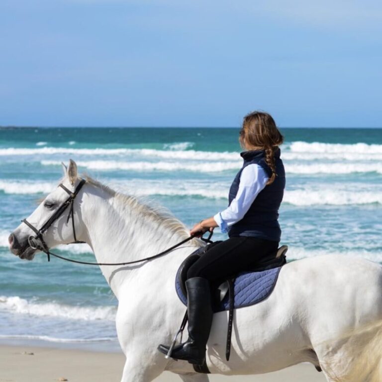 A woman on a white horse on a beach. She's looking to her right, at the sea. The sky is blue