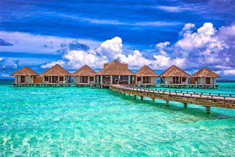 Huts on stilts over a turquoise sea. The sky is bright blue, there are a few cloud in the sky
